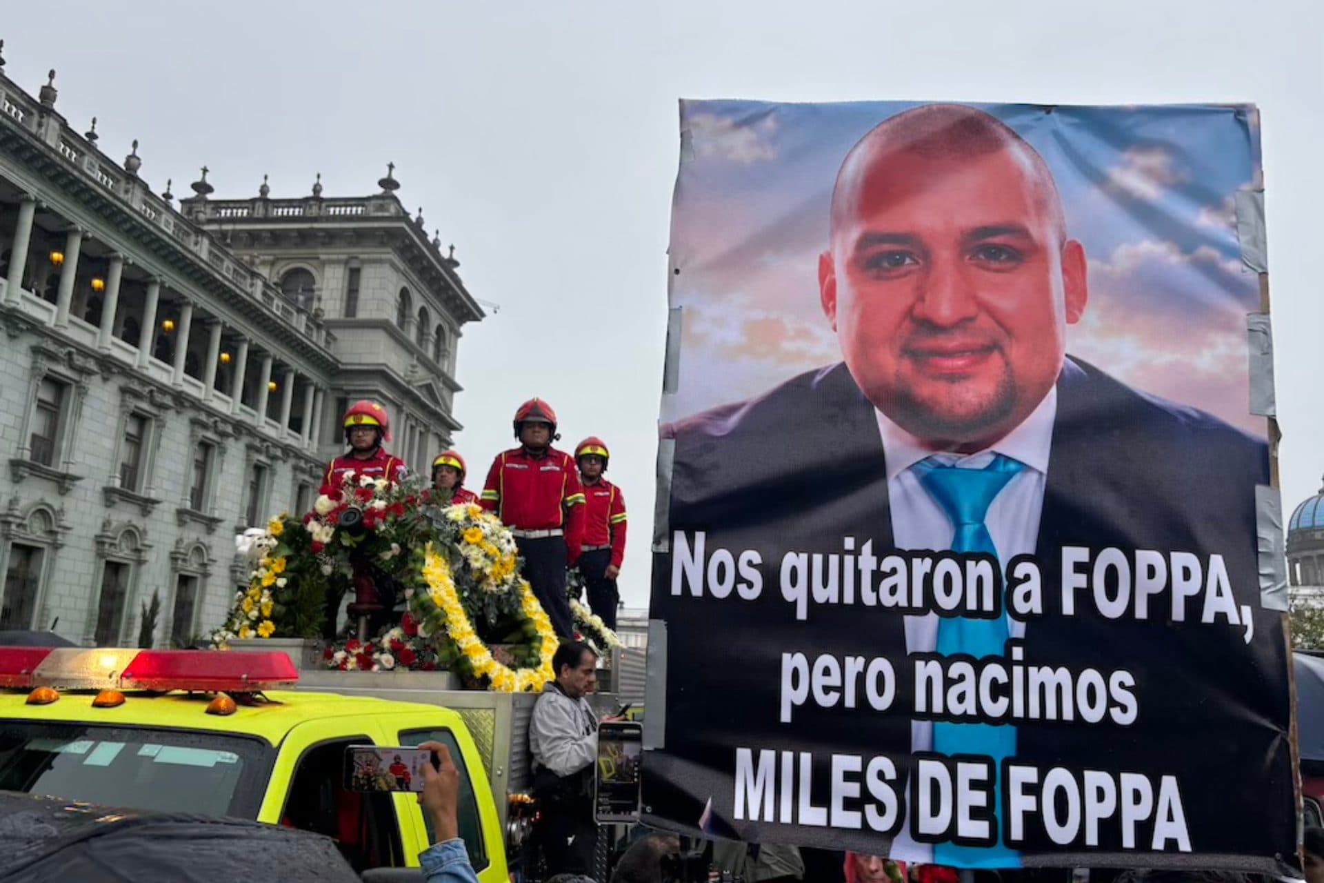 Bomberos voluntarios sobre un camión con arreglos florales frente al Palacio Nacional; al lado, una manta con el retrato de Juan Francisco Solórzano Foppa y la frase “Nos quitaron a Foppa, pero nacimos miles de Foppa”.