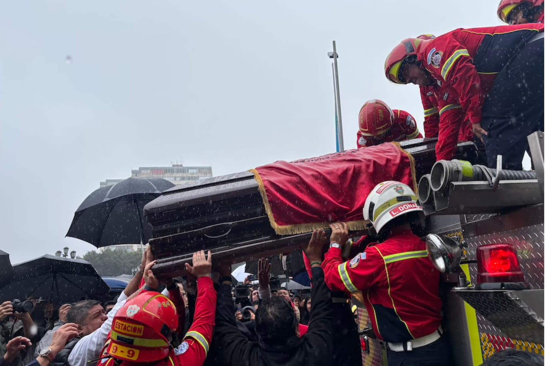 Bajo la lluvia, bomberos voluntarios suben a un camión el féretro de Solórzano Foppa, cubierto con una tela roja, mientras la gente alza las manos para ayudar.