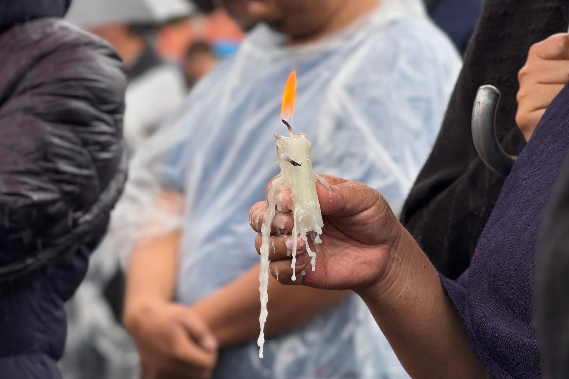 Una mano sostiene una vela encendida, cubierta de cera derretida, entre personas con capas para la lluvia.