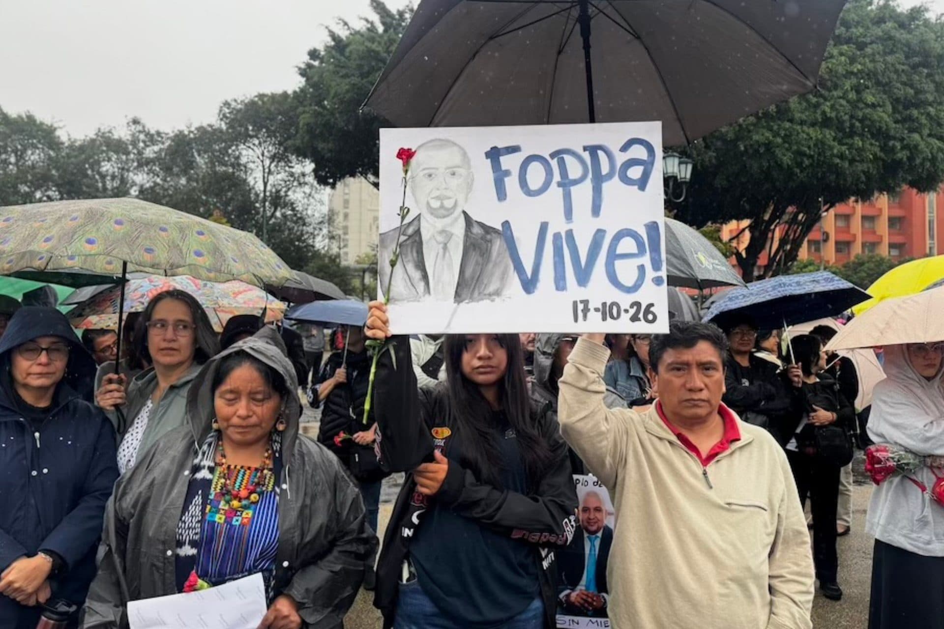 Una joven sostiene bajo un paraguas un cartel con un dibujo de Solórzano Foppa y la frase “Foppa vive! 17-10-26”, rodeada de otras personas en la plaza.