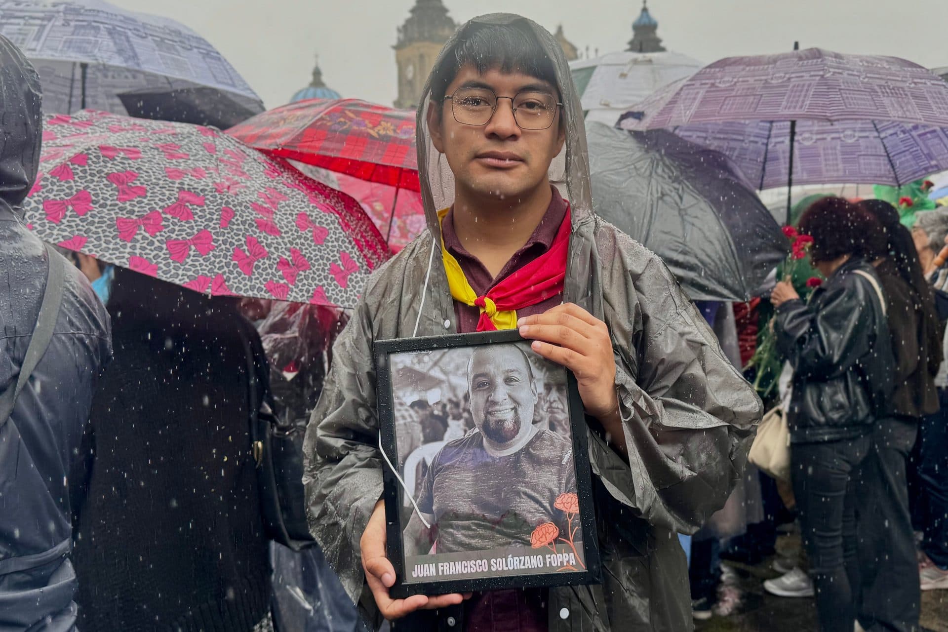 Un joven con capa para la lluvia y una pañoleta roja y amarilla sostiene un retrato enmarcado de Juan Francisco Solórzano Foppa en la plaza.