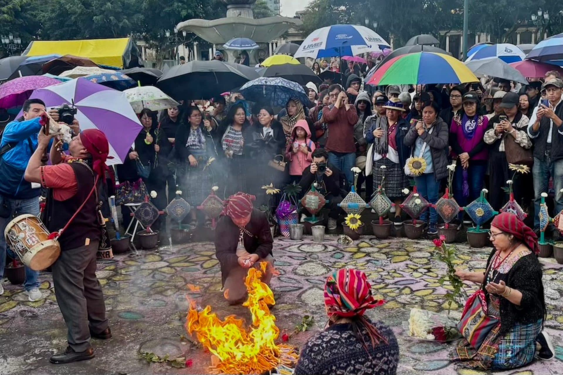 Guías espirituales mayas encienden el fuego ceremonial sobre un altar de flores y velas en la plaza, rodeados de personas con paraguas.