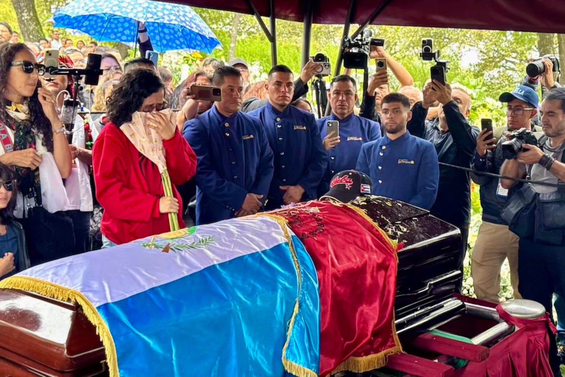 Junto al ataúd, cubierto con la bandera de Guatemala, una bandera roja y una gorra del equipo de béisbol de Cuba, una mujer se cubre el rostro con un pañuelo; detrás, hombres de uniforme azul y reporteros.